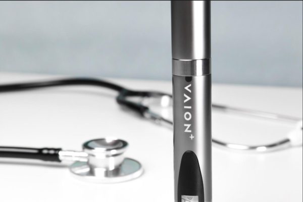 A VAION pen in focus is positioned upright on a flat surface, with a stethoscope blurred in the background. The VAION pen has a sleek metallic design with the word "VAION+" visible on it. The setting suggests a medical or health-related theme.