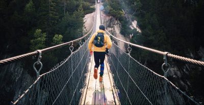 A person wearing a yellow jacket and a backpack is running across a suspension bridge, surrounded by dense forest. The bridge is high above a valley and stretches into the distance, with trees and rocky cliffs visible on either side.